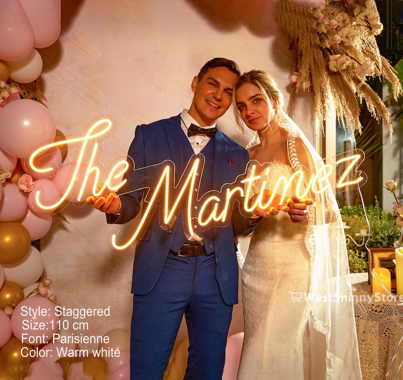 May include: A warm white neon sign that reads "The Martinez" in a cursive font. The sign is held by a couple, with the man in a blue suit and the woman in a wedding dress. The sign is 110 cm in size.