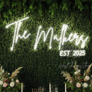 May include: A neon sign with the words "The Makers EST 2025" against a backdrop of green foliage. The sign is in a cursive font and glows brightly. Floral arrangements and candles are visible at the bottom.