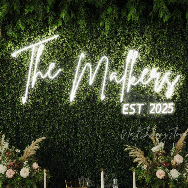 May include: A neon sign with the words "The Makers EST 2025" against a backdrop of green foliage. The sign is in a cursive font and glows brightly. Floral arrangements and candles are visible at the bottom.