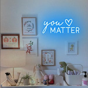 May include: A white changing table with a white changing mat, a white diaper pail, a white stuffed animal, a green dinosaur stuffed animal, a white lamp with a white shade, and a framed print with the text "You Matter" in blue neon lettering.