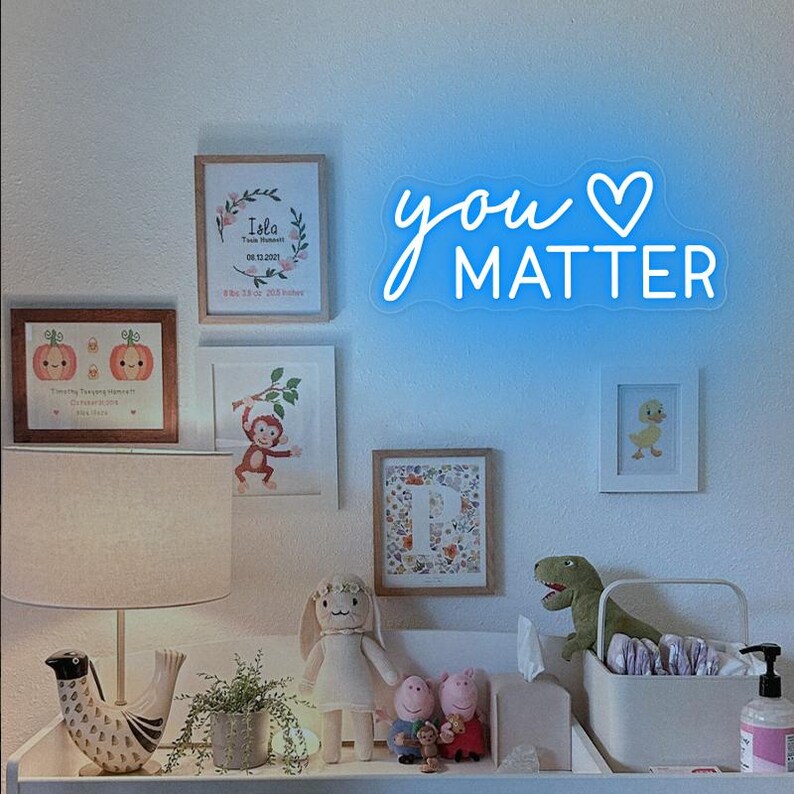 May include: A white changing table with a white changing mat, a white diaper pail, a white stuffed animal, a green dinosaur stuffed animal, a white lamp with a white shade, and a framed print with the text "You Matter" in blue neon lettering.