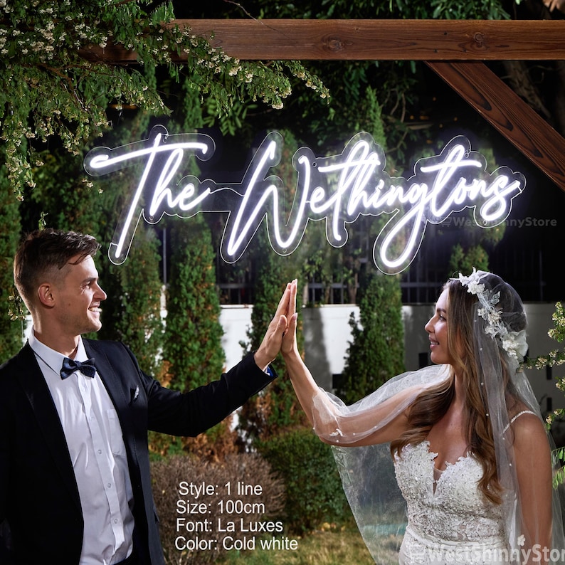 May include: A neon sign in the shape of the words "The Wethingtons" in a cursive font, lit with cold white light. The sign is 100cm in size. The sign is displayed outdoors with a couple in formal attire.
