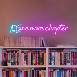 May include: A pink neon sign with the words "One more chapter" and a book icon. The sign is hanging on a wall above a bookshelf filled with books.