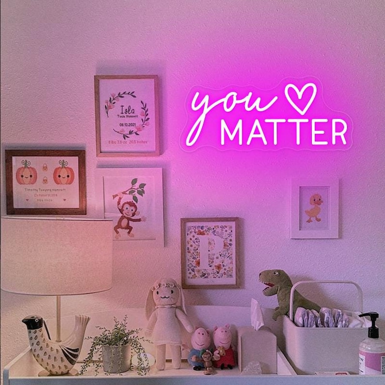 May include: Pink neon sign with the words "you" and a heart symbol, followed by the word "MATTER". The sign is hanging on a white wall with framed pictures and a lamp on a table below.