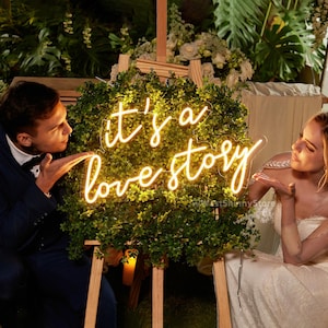 May include: A neon sign with the text "it's a love story" in a cursive font. The sign is lit up with a warm white glow and is placed in front of a green leafy backdrop. A bride and groom are standing on either side of the sign, smiling and looking at each other.