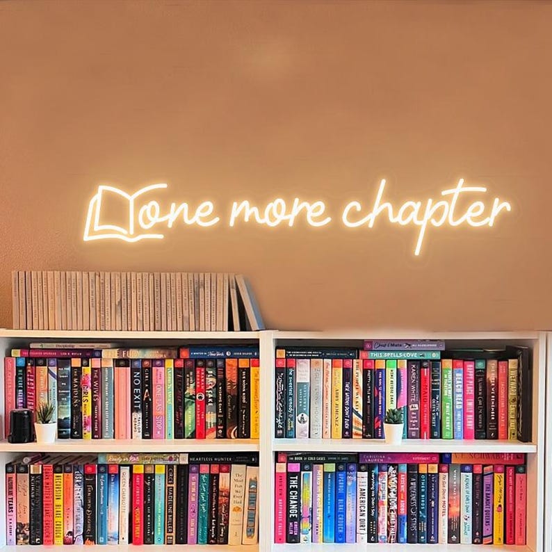 May include: A white neon sign with the text "Done more chapter" and a stylized image of an open book. The sign is mounted on a wall above a bookshelf filled with books.