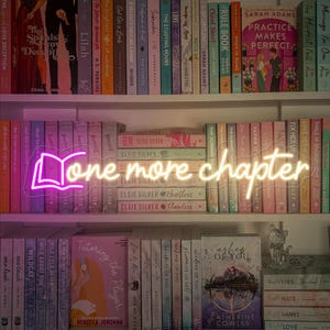 May include: A neon sign with a pink book shape and the words "one more chapter" in yellow. The sign is on a shelf with many colorful books.