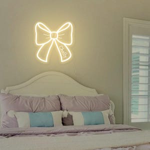May include: A white neon sign shaped like a bow with the name "Chelly" written on it. The sign is hanging on a wall above a white headboard with pillows.