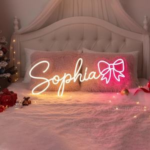May include: A neon sign spelling "Sophia" in cursive script, with a bow graphic. The sign is lit with a warm white and pink glow, resting on a bed with pink pillows and a fluffy blanket. A Christmas tree and gifts are in the background.