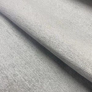 May include: A light gray fabric with a textured, slightly nubby surface. The material is folded over itself, creating a diagonal crease that highlights the consistent, dense weave and the matte, neutral tone of the textile throughout the piece.