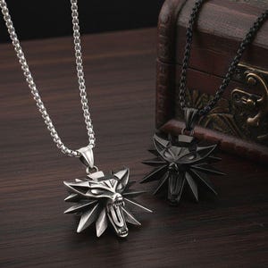 May include: Two wolf head pendant necklaces. One is silver-toned, the other is black. Both pendants have a detailed wolf head design with sharp teeth and a spiky frame. The necklaces are displayed on a dark wooden surface.