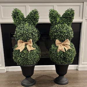 Peut inclure: Deux topiaires décoratifs en forme de lapin de Pâques. Chaque topiaire est faite de feuillage vert en forme de lapin avec un nœud en toile de jute. Les lapins sont dans des jardinières noires en forme d'urne. L'arrière-plan comprend une cheminée et des boiseries blanches.