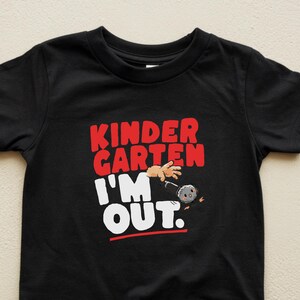 May include: Black toddler t-shirt with the text "KINDER GARTEN I'M OUT." in red and white. The graphic includes a cartoon hand and a bowling ball. A red line underlines "OUT."