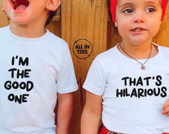 May include: Two children wearing white t-shirts with black text. One shirt says "I'M THE GOOD ONE", the other says "THAT'S HILARIOUS". The child on the right has a red headband and a red and black plaid shirt tied around the waist.