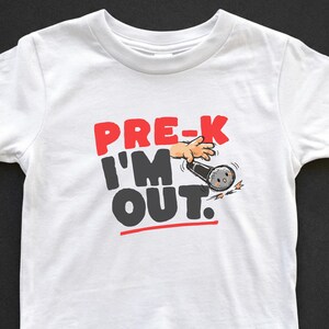 May include: White t-shirt with the text "PRE-K I'M OUT" in red and black, with a cartoon illustration of a hand holding a microphone. The shirt is designed for children.
