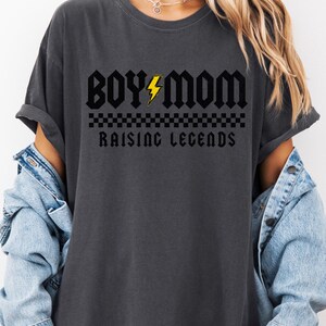 Peut inclure: T-shirt gris fonc&eacute; avec l'inscription "BOY MOM" en police gothique, un &eacute;clair jaune et les mots "RAISING LEGENDS" en dessous. Un motif &agrave; carreaux est plac&eacute; entre les mots. Le t-shirt est port&eacute; avec une veste en jean.