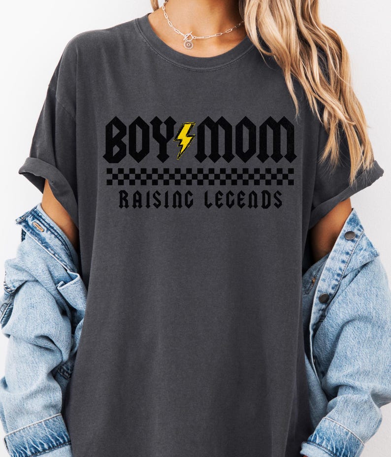 Peut inclure: T-shirt gris fonc&eacute; avec l'inscription "BOY MOM" en police gothique, un &eacute;clair jaune et les mots "RAISING LEGENDS" en dessous. Un motif &agrave; carreaux est plac&eacute; entre les mots. Le t-shirt est port&eacute; avec une veste en jean.