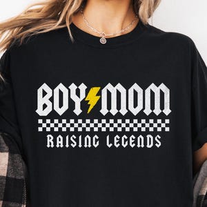 Peut inclure: T-shirt noir avec l'inscription blanche "BOY MOM", un &eacute;clair jaune et la phrase "RAISING LEGENDS" dans une police de style rock and roll. Un motif &agrave; carreaux est entre les phrases.