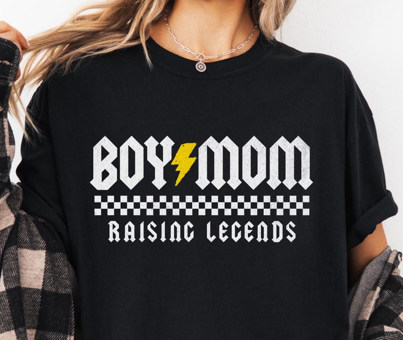 Peut inclure: T-shirt noir avec l'inscription blanche "BOY MOM", un &eacute;clair jaune et la phrase "RAISING LEGENDS" dans une police de style rock and roll. Un motif &agrave; carreaux est entre les phrases.