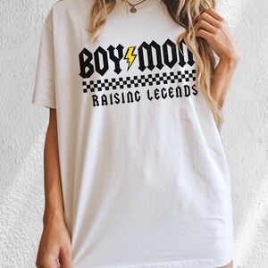 Peut inclure: T-shirt blanc avec l'inscription "BOY MOM RAISING LEGENDS" en police gothique noire. Un &eacute;clair jaune est plac&eacute; entre les mots "BOY" et "MOM". Un motif &agrave; carreaux noir et blanc se trouve en dessous.