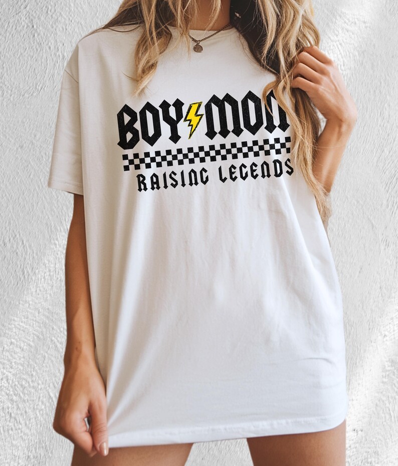 Peut inclure: T-shirt blanc avec l'inscription "BOY MOM RAISING LEGENDS" en police gothique noire. Un &eacute;clair jaune est plac&eacute; entre les mots "BOY" et "MOM". Un motif &agrave; carreaux noir et blanc se trouve en dessous.