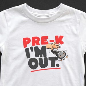 May include: White toddler t-shirt with the text "PRE-K I'M OUT" in red and black. A cartoon hand is holding a microphone, with a red line under the word "OUT". The shirt is made of a soft, comfortable material.