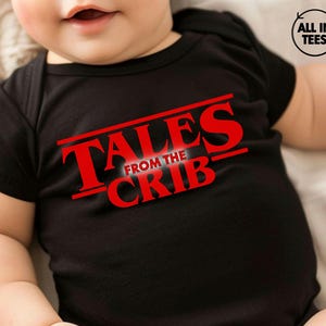 May include: Black baby onesie with the red text "TALES FROM THE CRIB" in a bold, angled font. The onesie is short-sleeved and appears to be made of soft material. The image also includes a small logo in the upper right corner.