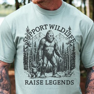 May include: A light green t-shirt featuring a black and white illustration of a Bigfoot with a smaller Bigfoot holding its hand. The text reads "SUPPORT WILDLIFE" above and "RAISE LEGENDS" below the image.