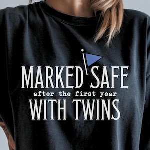 May include: Black t-shirt with white text that reads "MARKED SAFE after the first year WITH TWINS." A blue flag graphic is above the word "SAFE." The shirt is a dark color and appears to be made of soft material.