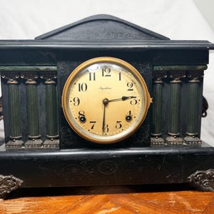 May include: Antique black mantel clock with a gold-toned clock face. The clock features decorative green columns and ornate detailing on the base and top. The clock face has black numbers and hands, and the brand name "Ingraham" is visible.