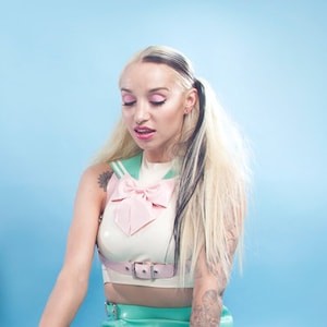 Puede incluir: Una mujer con un top blanco y rosa con un lazo, una falda verde y botas blancas hasta el muslo se sienta sobre un fondo azul. Está jugando con un tren de juguete y dos ponis de juguete.