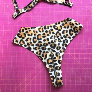 Może przedstawiać: Zestaw bielizny w panterkę składający się z topu z uprzężą i majtek z wysokim stanem. Uprząż ma czarne paski i złote okucia. Majtki mają zapięcie na zamek. Zestaw wykonany jest z tkaniny z czarno-brązowo-beżowym zwierzęcym nadrukiem.