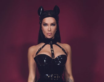 May include: A woman wearing a black latex bodysuit with gold hardware and a black cat ear headband. The bodysuit features a corset-style top with a plunging neckline and a zipper down the front. The legs of the bodysuit are fitted and have straps with buckles around the thighs.