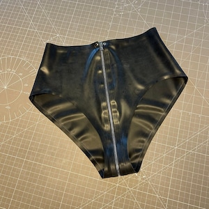 Peut inclure: Culotte taille haute noire en matière brillante, avec une fermeture éclair sur toute la longueur à l'avant. Le vêtement est présenté sur un tapis de coupe gris.