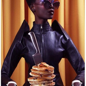 Pu&ograve; includere: Una persona con una giacca di pelle nera e guanti tiene un coltello sopra una pila di pancake ricoperti di sciroppo su un piatto bianco.