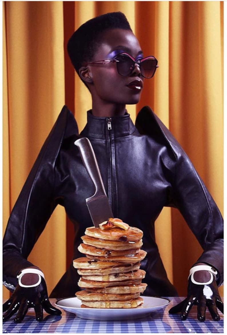 Pu&ograve; includere: Una persona con una giacca di pelle nera e guanti tiene un coltello sopra una pila di pancake ricoperti di sciroppo su un piatto bianco.
