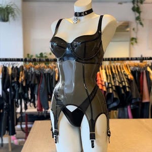 Könnte beinhalten: Schwarzes Latex-Dessous-Set auf einer Schaufensterpuppe. Das Set besteht aus einem BH, einem Korsett und einem Strumpfhalter. Ein schwarzes Halsband mit einem silbernen Herzanhänger vervollständigt den Look. Die Dessous sind glänzend und figurbetont.