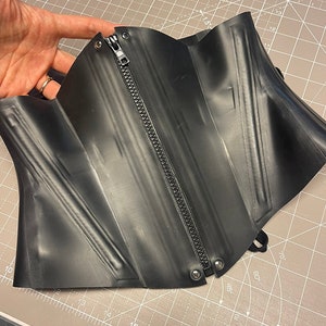 May include: Black rubber corset with a zipper down the front and silver grommets. The corset is lying on a gray cutting mat.