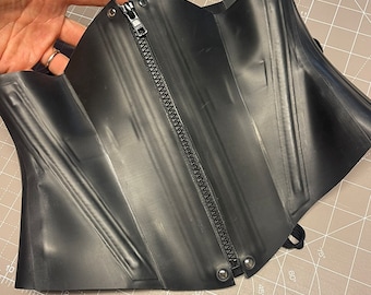 May include: Black rubber corset with a zipper down the front and silver grommets. The corset is lying on a gray cutting mat.