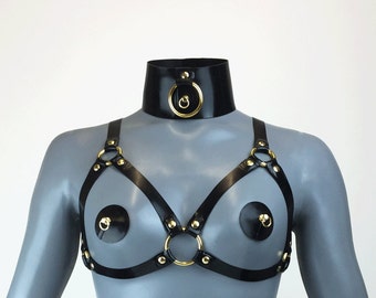May include: Black leather harness with gold rings and studs. The harness has two straps that go over the shoulders and connect in the front with a large gold ring. There are two smaller gold rings on each strap, and two black nipple pasties with gold rings.