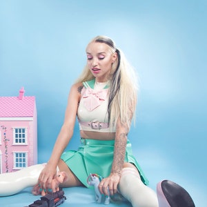 May include: A woman wearing a white and pink sailor-style top, a green skirt, and white thigh-high boots sits on a blue background. She is playing with a toy train, a pink pony, and a blue unicorn.