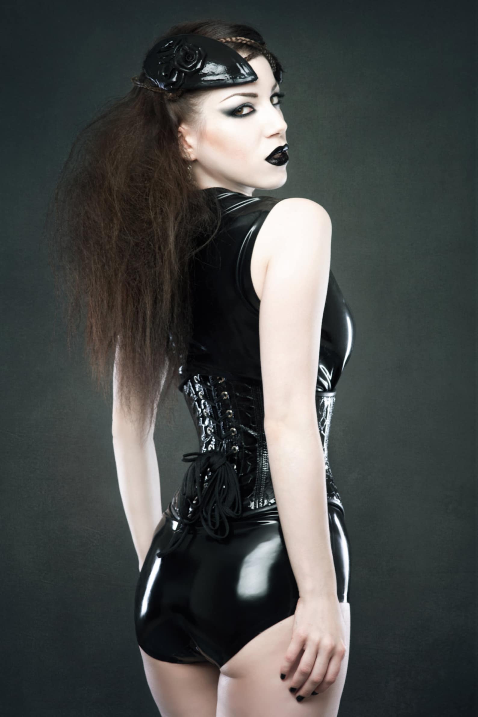 Back view of a gothic model in a black latex bodysuit with a corset waist, teased hair, glossy black headpiece, and dramatic eye makeup.