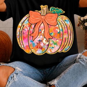 Può includere: Una t-shirt nera a maniche corte con una grande stampa di una zucca. La zucca presenta un vivace motivo floreale multicolore, un fiocco arancione a tinta unita legato sulla parte superiore e un contorno glitterato dorato. Il design include una foglia verde sul picciolo.