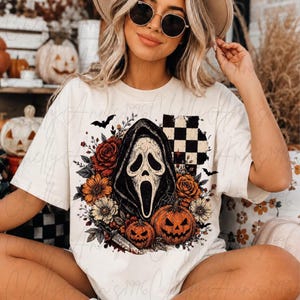 May include: A cream-colored short-sleeve t-shirt featuring a graphic print of a ghost-like mask in a dark hood, surrounded by orange and red flowers, two jack-o'-lanterns, a small knife, a black bat, and a black and white checkered square pattern.