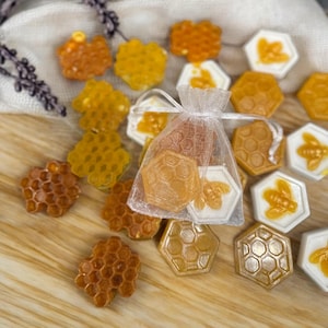 May include: A collection of hexagon-shaped soaps in various shades of yellow and brown, resembling honeycombs. Some soaps feature bee designs. A small, sheer bag contains several soaps. The soaps are arranged on a wooden surface.