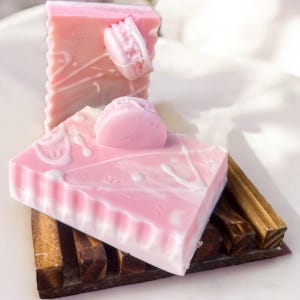 Puede incluir: Dos barras de jabón de color rosa y blanco con un diseño de espiral decorativo. Las barras de jabón están sobre un jabonera de madera.