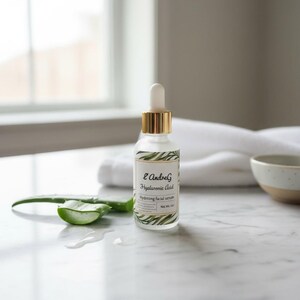 May include: A clear glass bottle of L'AndreG Hyaluronic Acid Hydrating facial serum with a gold-colored cap and white dropper. The label has green leaf accents. A sliced aloe vera leaf and water droplets are next to the bottle. The background includes a white towel and a small bowl.