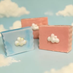 May include: Three rectangular soaps in pastel colors: light blue, and two shades of pink. Each soap has a white cloud-shaped embellishment. The soaps are arranged on a light blue surface with a cloudy sky background.