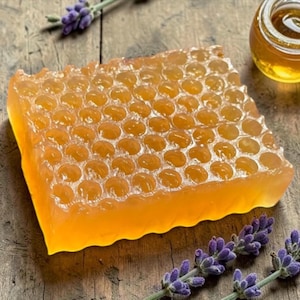 Puede incluir: Primer plano de una pastilla de jabón con forma de panal de abeja de color amarillo dorado. El jabón está rodeado de ramitas de lavanda y un pequeño tarro de miel, todo sobre una superficie de madera rústica. El diseño del jabón imita un panal natural.