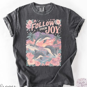 May include: A charcoal gray short-sleeve t-shirt featuring a graphic print of three dolphins leaping from ocean waves. The design includes pink hibiscus flowers, palm trees, a sunset, and the text "FOLLOW YOUR JOY" in a light pink font.